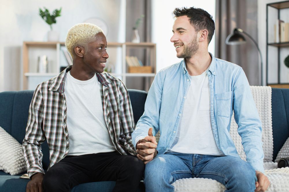Interracial Gay couple sits in therapy office smiling and clasping hands.