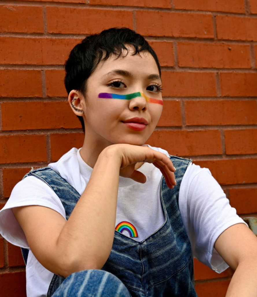 Content woman with rainbow face paint and overalls poses for a photo against a brick wall, confient after receiving individual therapy at Brickel & Associates