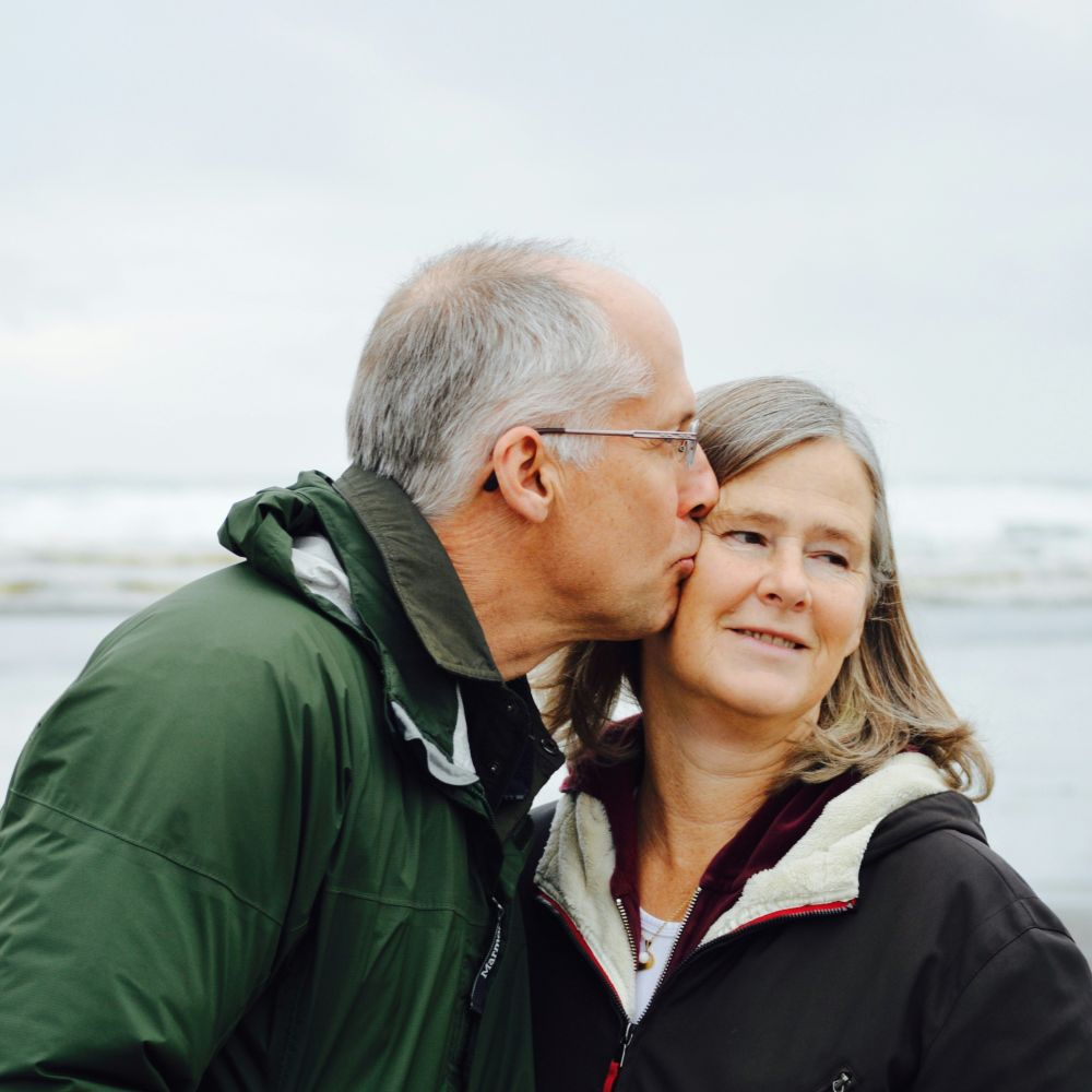 An older man kisses his wife on the beach after finding comfort and understanding in relational therapy.