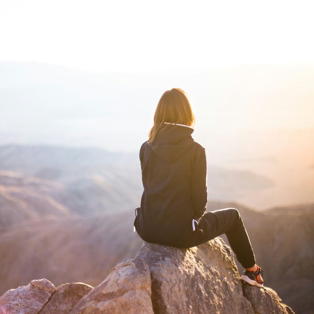 A strong and confident women sits at the top of a mountain overlooking the sunset.